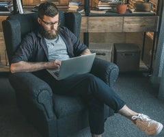 bearded man in eyeglasses using laptop while sitting in armchair at home