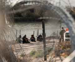 Migrantes esperan con miembros de la Guardia Nacional de Texas a ser procesados por la patrulla fronteriza a orillas del río Grande en Eagle Pass.