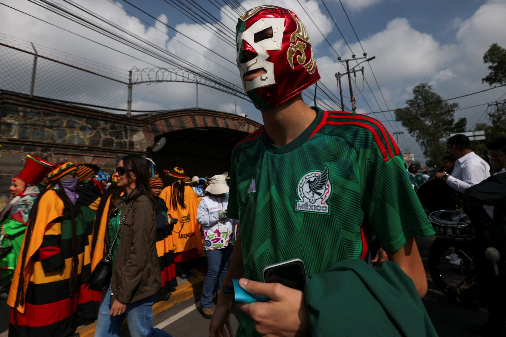 Un hombre con una máscara de luchador camina entre una multitud de aficionados que pasan por un control policial, donde verifican sus entradas